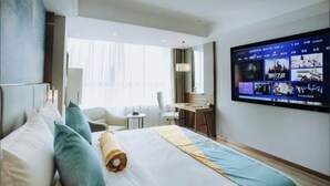 Quarto de hotel com uma cama grande, uma escrivaninha, uma cadeira e uma TV exibindo títulos de jogos.