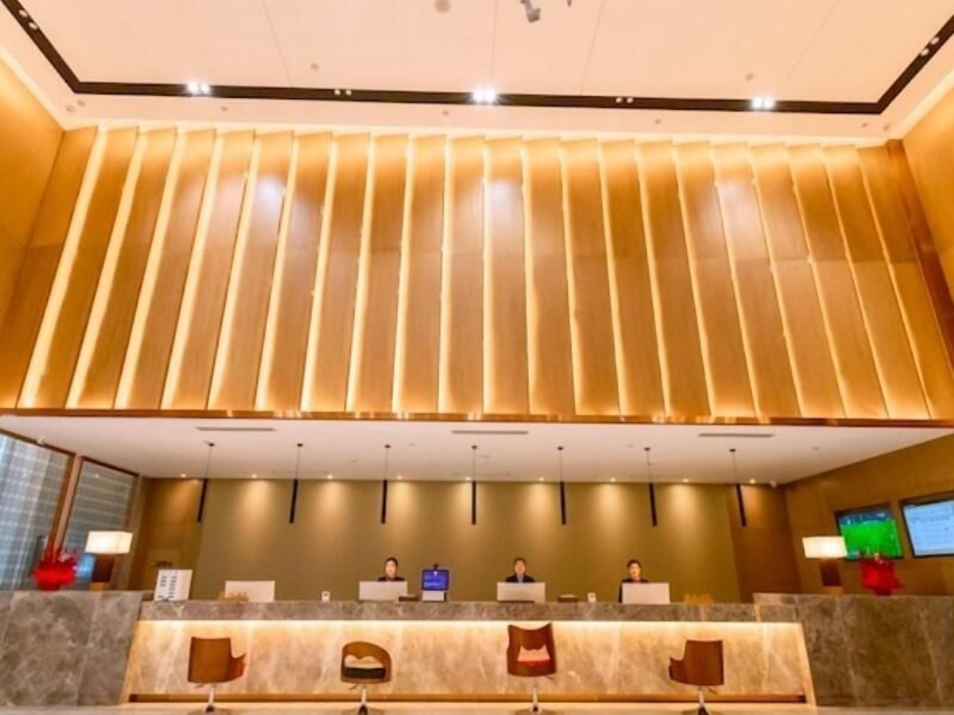 Ein Hotel-Empfangsbereich mit modernem Design, der eine Rezeption, mehrere Stühle und einen großen Bildschirm mit Informationen umfasst.