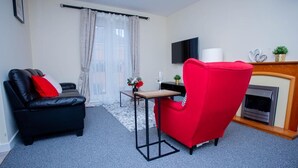Ein Wohnzimmer mit schwarzem Sofa, einem roten Sessel, einem kleinen Tisch mit Blumenvase, einem flachbildfernseher an der Wand und einem Kamin.