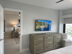 A modern bedroom with a large TV displaying a scenic view, a wooden dresser, and a view of the outdoors. - Beautifully remodeled condo offering breathtaking Gulf views right on the beach. (Marco Island)