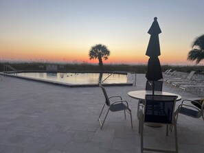 A poolside area with lounge chairs and an umbrella, overlooking a body of water and a sunset.