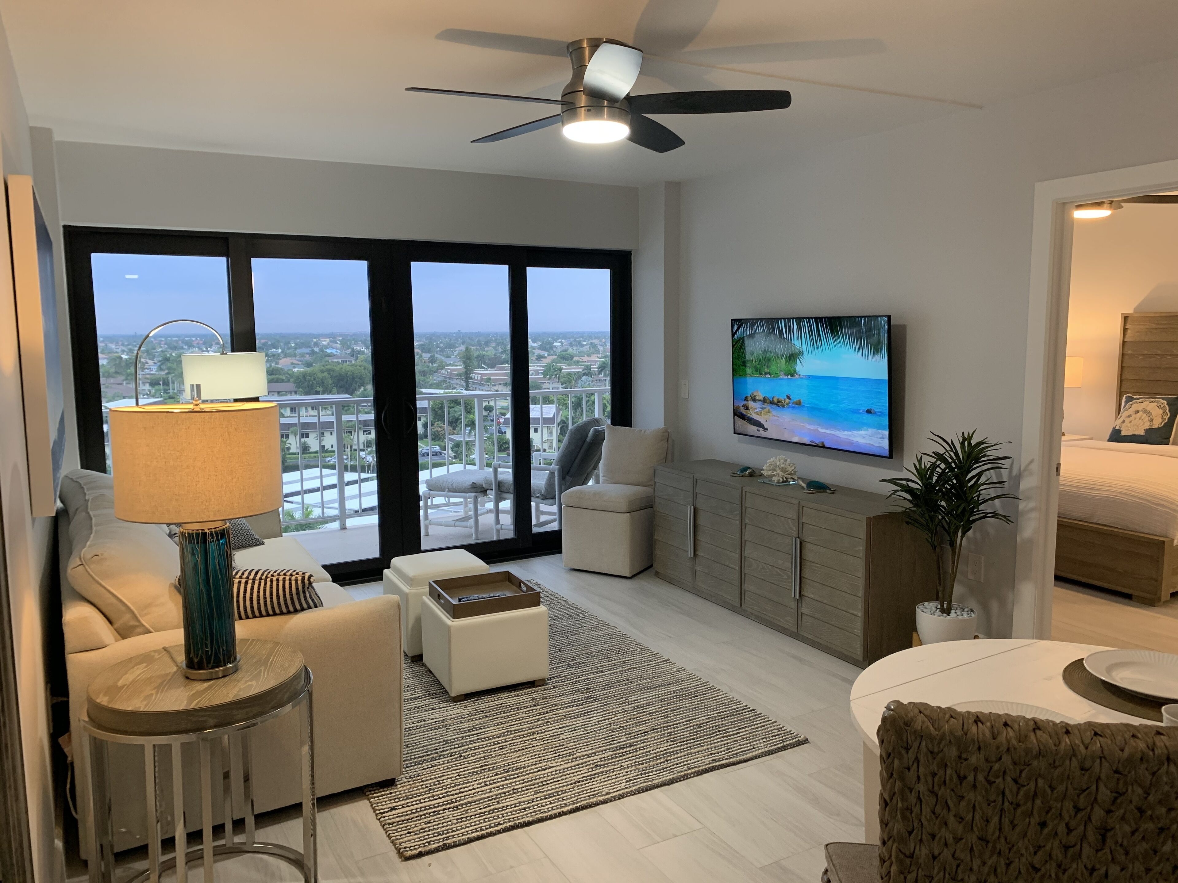 A modern living room with a large TV, comfortable seating, a balcony with a view, and a ceiling fan.