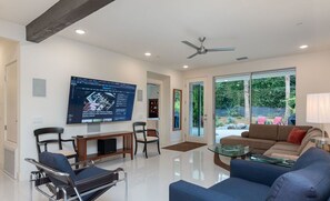 A spacious living room with a large TV, comfortable seating, and a glass door leading to a garden.