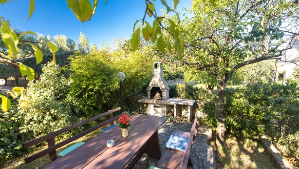 A stone barbecue area with a chimney, surrounded by lush greenery and a wooden table with a vase of flowers.