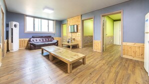 Una habitación con piso de madera, un sofá de cuero, una mesa de madera, un televisor en la pared y una puerta que da a otra habitación.