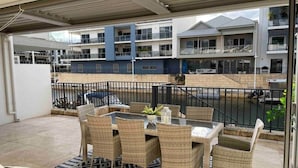 Een overdekt terras met rieten meubilair en een vloerkleed met patroon, met uitzicht op een zwembad en een meerlaags gebouw met balkons.