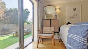Une pièce avec une grande fenêtre, une chaise en bois, un lit recouvert d’une couverture bleue et une œuvre d’art encadrée accrochée au mur.