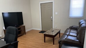 Uma sala de estar com televisão, sofá de couro, mesa de centro e uma porta.