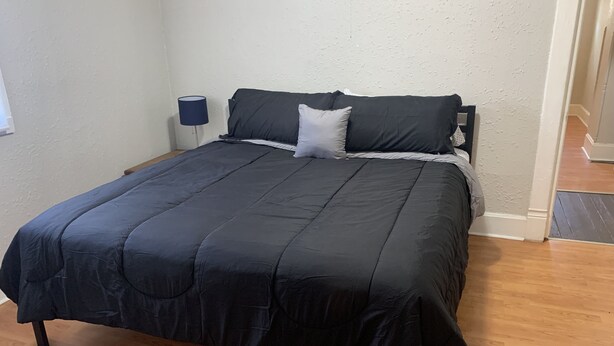 Cama bem arrumada com colcha escura e almofadas, luminária de cabeceira e assoalho de madeira.