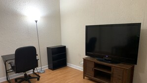 Um quarto com piso de madeira, uma cadeira de escritório preta, um abajur, uma TV em um suporte de madeira e uma estante preta.