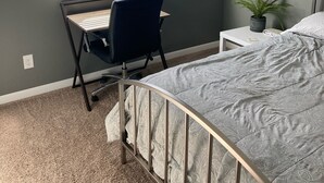 Ett sovrum med en säng, ett skrivbord med en stol, ett fönster med persienner, en krukväxt och ett nattduksbord.