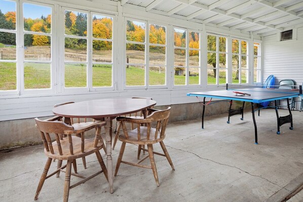 A covered patio with a round wooden table and chairs, a ping pong table, and large windows overlooking a grassy field with autumn trees.