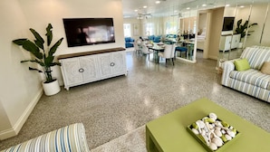 Uma sala de estar moderna com uma TV de tela plana, uma área de jantar e uma cozinha visíveis através de um painel de vidro.