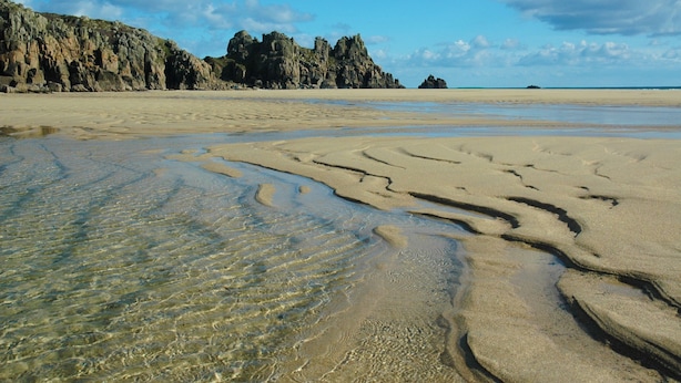 A beach with clear shallow waters and rocky formations in the background.