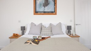 A neatly made bed with a book, a bowl of fruit, and a wooden tray. There are two bedside tables with lamps and a framed picture above the bed.