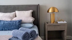 Een netjes opgemaakt bed met opgerolde handdoeken, een bedlampje en een klein houten nachtkastje met een boek en decoratieve spullen.