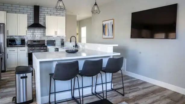 Een moderne keuken met witte kasten, een centraal eiland met barstoelen, een roestvrijstalen prullenbak en hanglampen.
