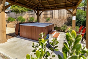 Relax in a serene outdoor oasis with a hot tub surrounded by lush greenery and a shaded pergola.