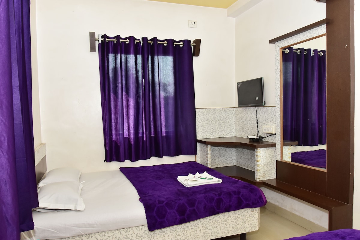A room with a bed, purple curtains, a small desk with a mirror, and a television.