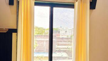A room with a bed, an air conditioner, a window with curtains, and a view of buildings outside.