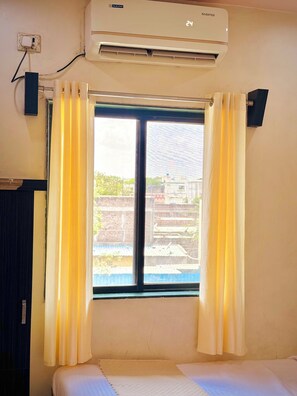 A room with a bed, an air conditioner, a window with curtains, and a view of buildings outside. - Hotel Temple Sai (Rahata)