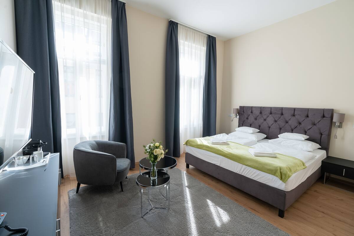 A modern hotel room with a bed, a desk, a chair, and a small table with a vase of flowers.