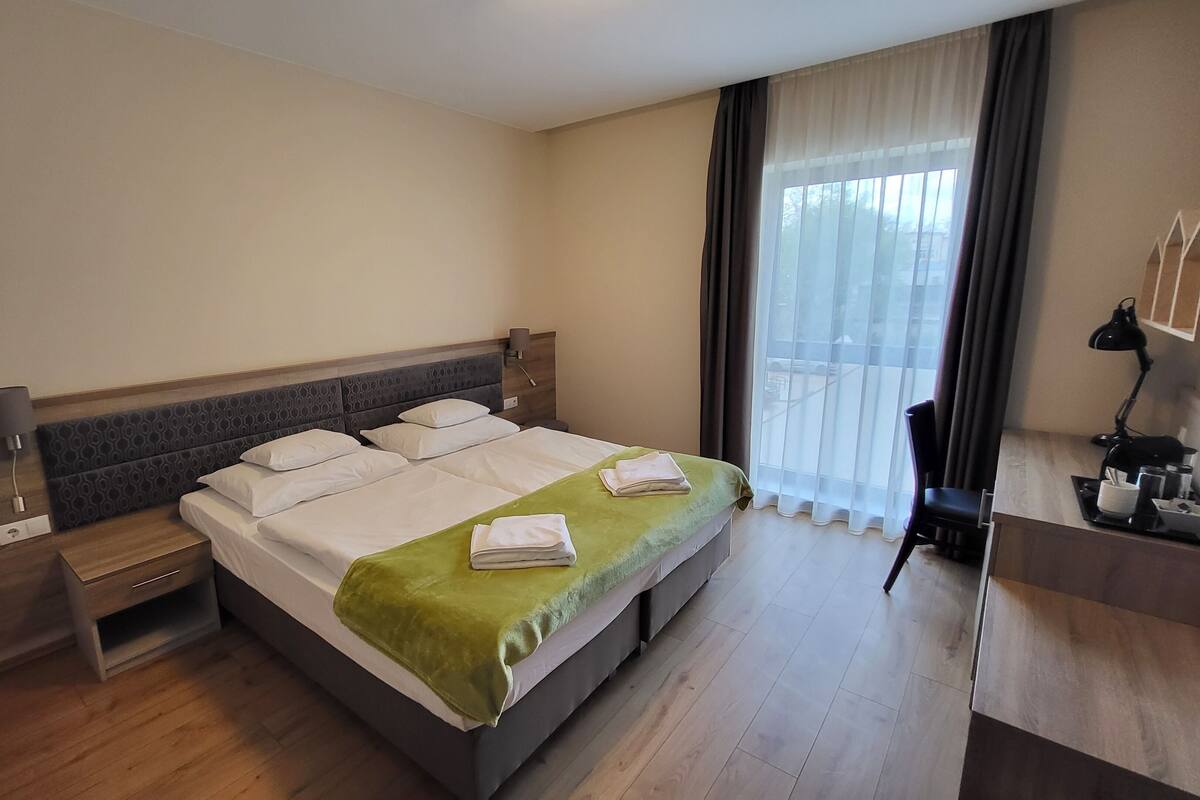 A hotel room with a bed, bedside table, lamp, and a view of the outside through a window.