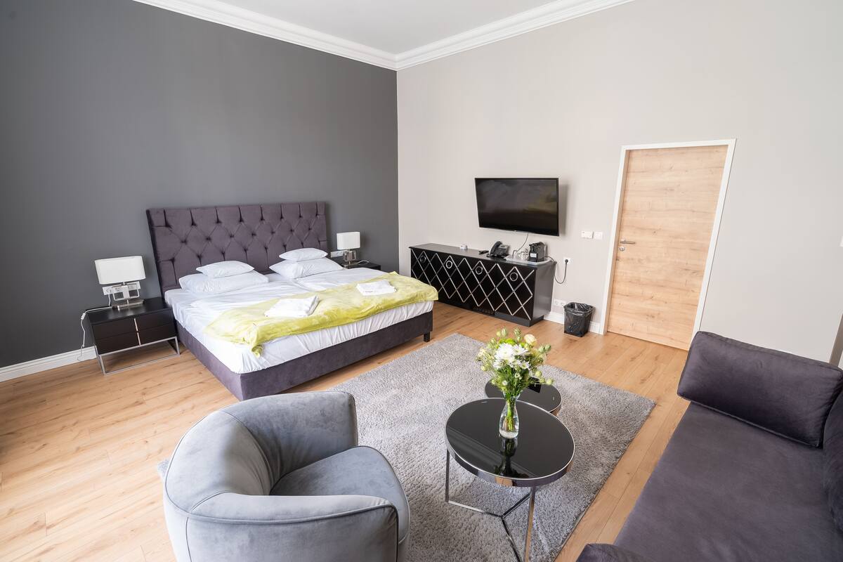A modern hotel room with a large bed, a flat-screen TV, a sofa, a small table with a vase of flowers, and a wooden door.