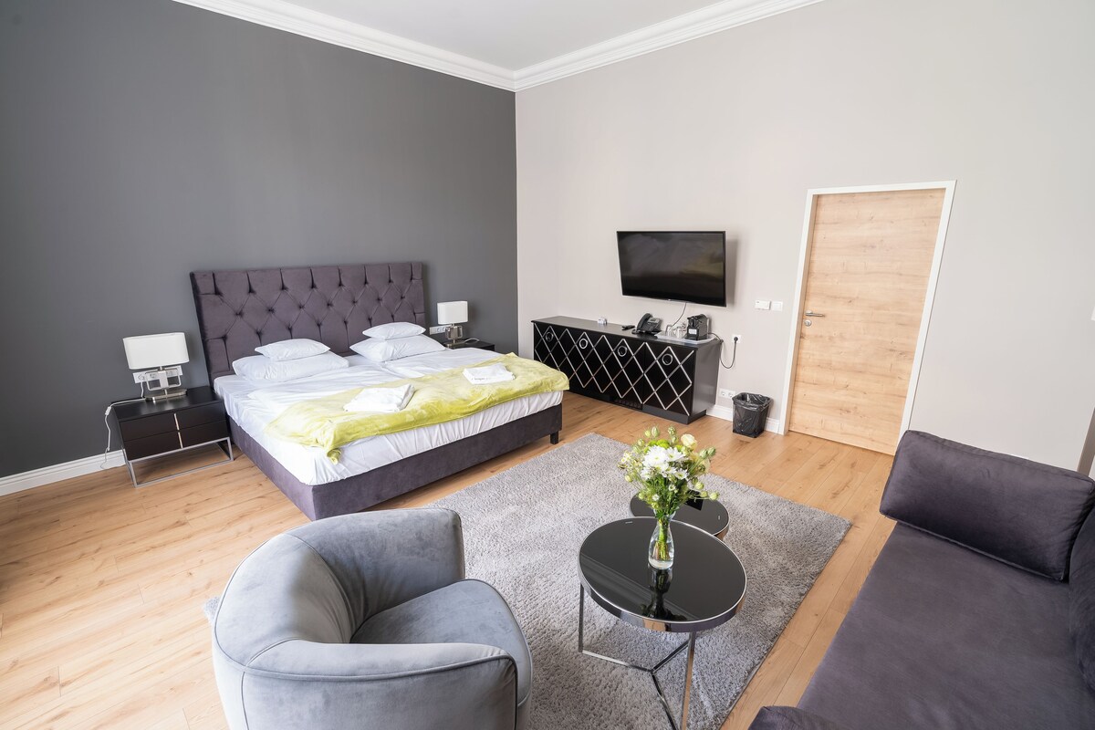 A modern hotel room with a large bed, a flat-screen TV, a sofa, a small table with a vase of flowers, and a wooden door.