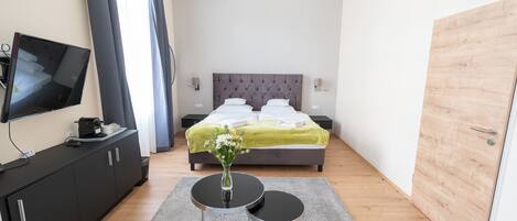 A modern hotel room with a bed, a TV on a black cabinet, a small table with a vase, and a wooden door.