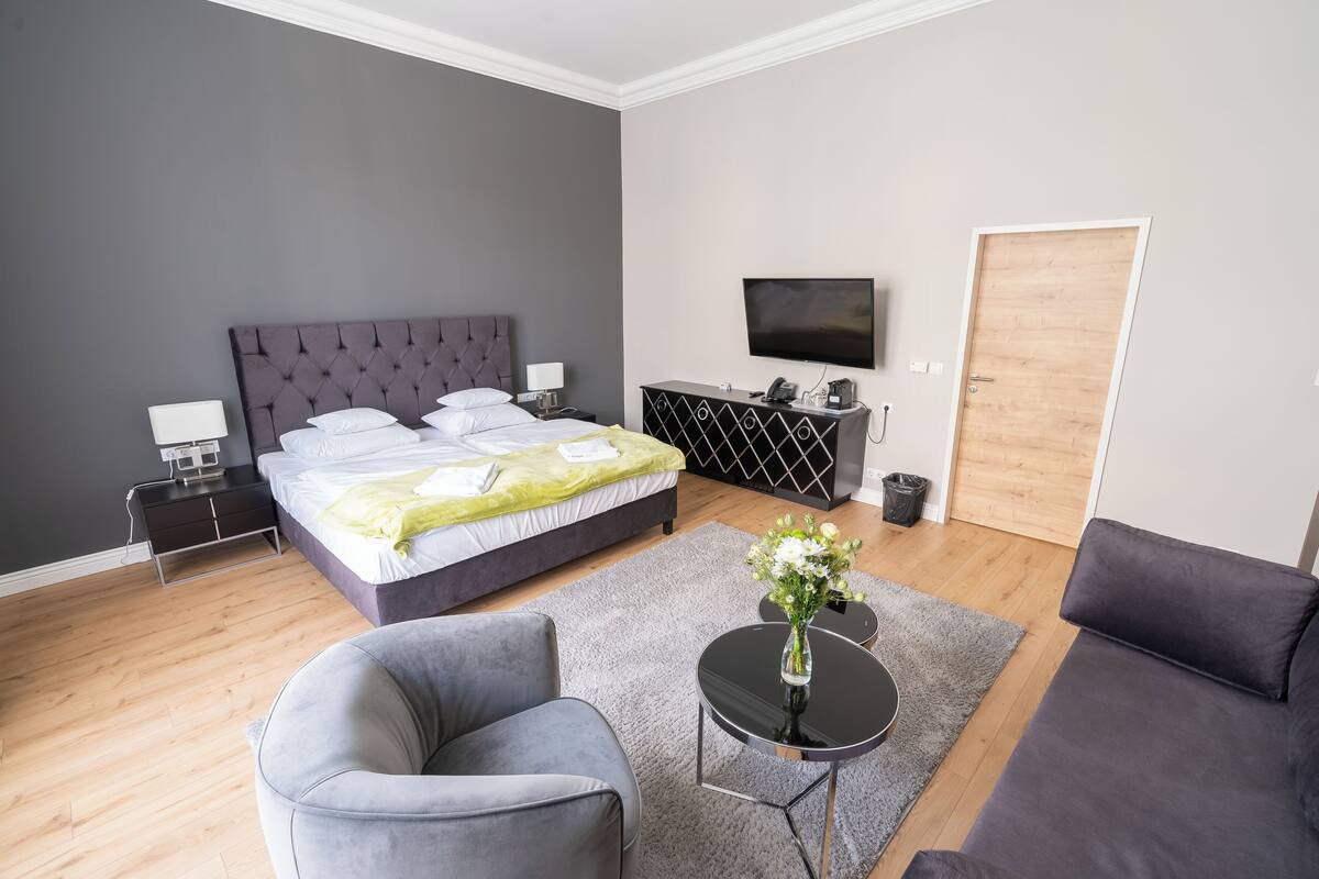 A modern hotel room with a large bed, a flat-screen TV, a sofa, a small table with a vase of flowers, and a wooden door.