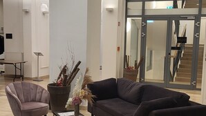 Un hall d’entrée spacieux au design moderne, avec un canapé aux couleurs sombres, un fauteuil rose et une table basse agrémentée d’objets décoratifs.