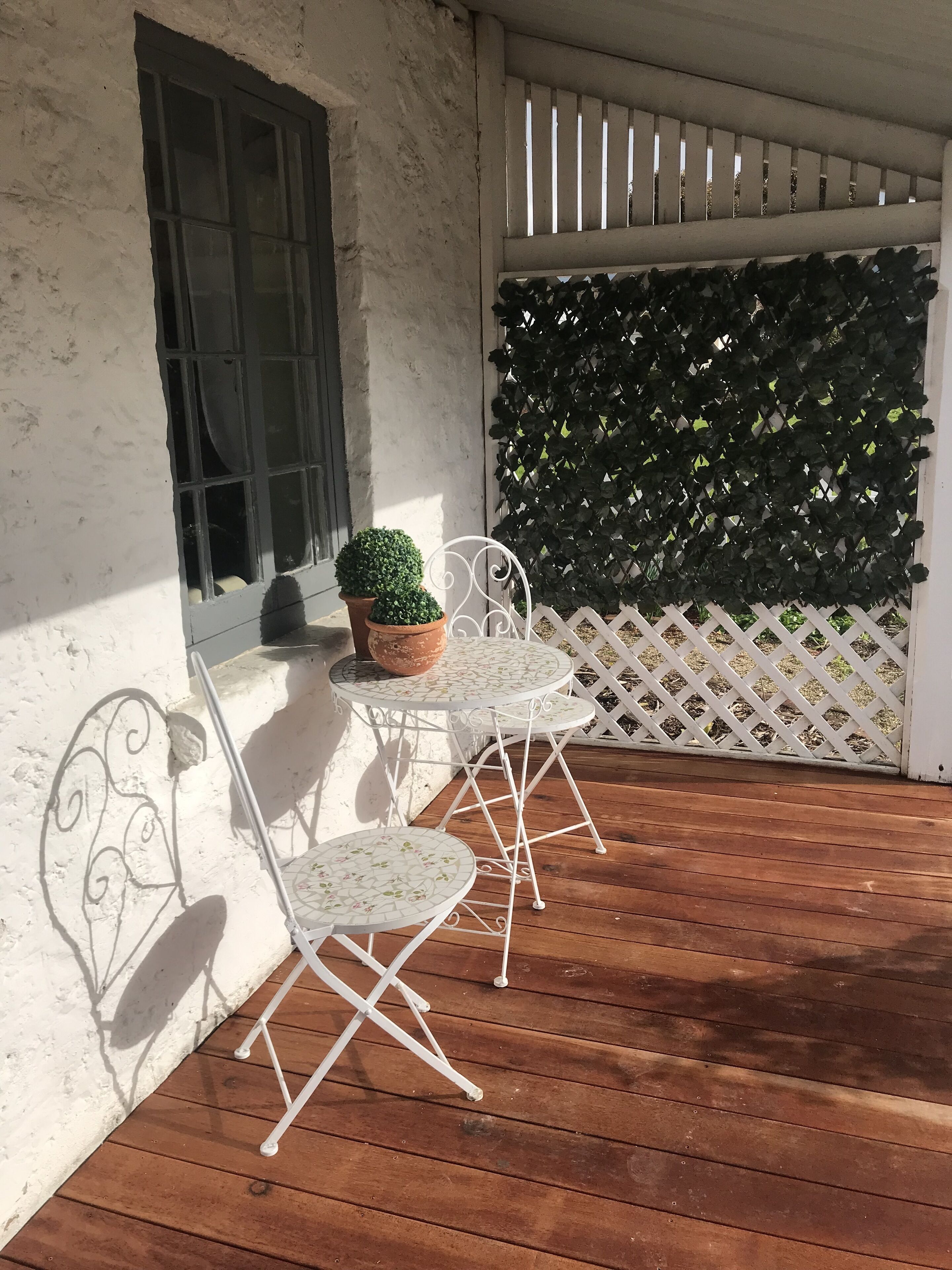 Una terraza de madera con dos sillas blancas de metal y una mesita, una ventana con cortina, y un muro de celosía con follaje.