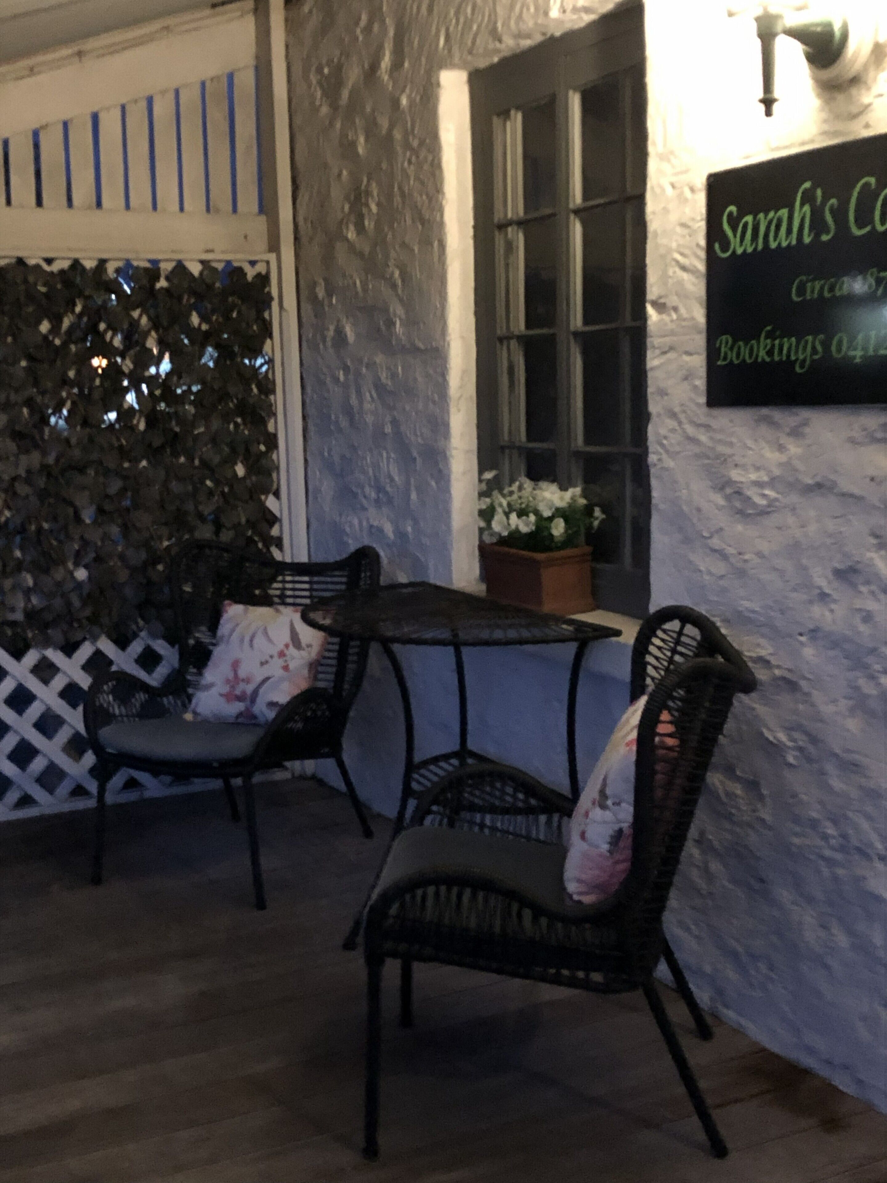 An outdoor seating area with wicker chairs and a table, a white picket fence, and a wall-mounted sign with text.