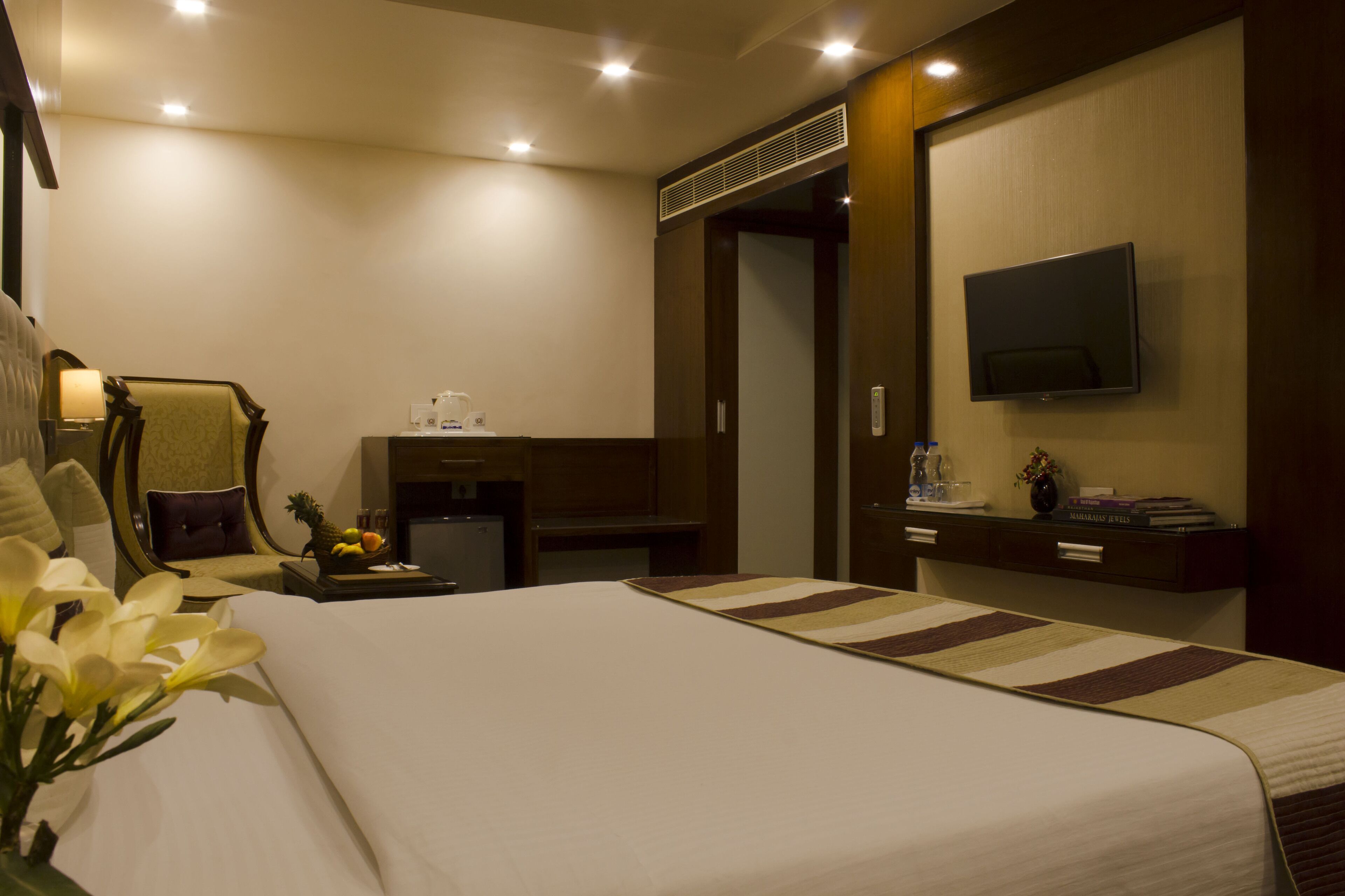 Une chambre d’hôtel avec un grand lit, un bureau avec un ordinateur, une chaise, un téléviseur fixé au mur et une petite table avec une lampe et un panier de fruits.