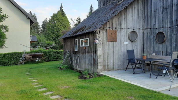 A rustic wooden building with a patio area featuring a table and chairs, a barbecue grill, and a string of lights. - Guesthouse Buberl, near Legoland and Skylinepark (Krumbach (Schwaben))