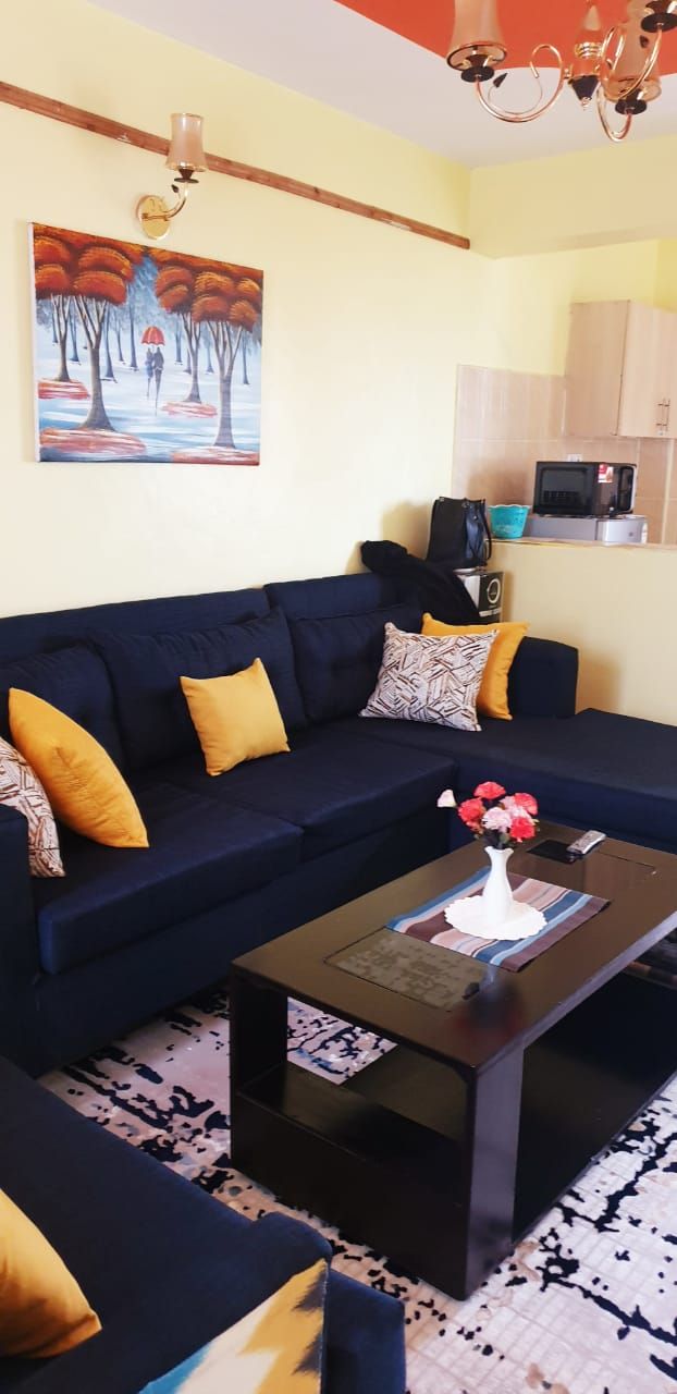 Un salón con un sofá azul oscuro, cojines amarillos, una mesita de centro con un jarrón de flores y un cuadro en la pared.