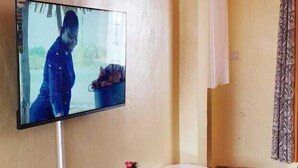 A living room with a flat-screen TV mounted on the wall, a black entertainment unit, a small white table, and a patterned rug. - Stay.Plus TRM Apartments (Nairobi)