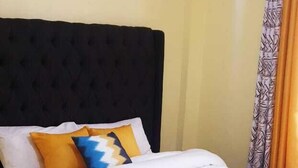 A neatly made bed with a dark headboard, white and yellow bedding, and a patterned rug on the floor. - Stay.Plus TRM Apartments (Nairobi)