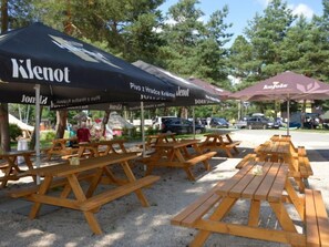 Picknicktische und Sonnenschirme im Freien mit 'Klenot'-Branding, in einem Parkplatz mit Autos und Bäumen im Hintergrund.