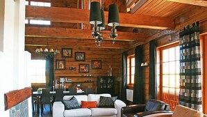 Un acogedor interior de cabaña de madera con un sofá blanco, una mesa de centro negra y un sillón de cuero. Se observan vigas de madera en el techo y una chimenea al fondo.