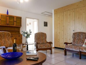Ein Wohnzimmer mit einer Holzkommode, zwei gemusterten Sesseln, einem runden Couchtisch, einer Blumenvase und einer Weinflasche.