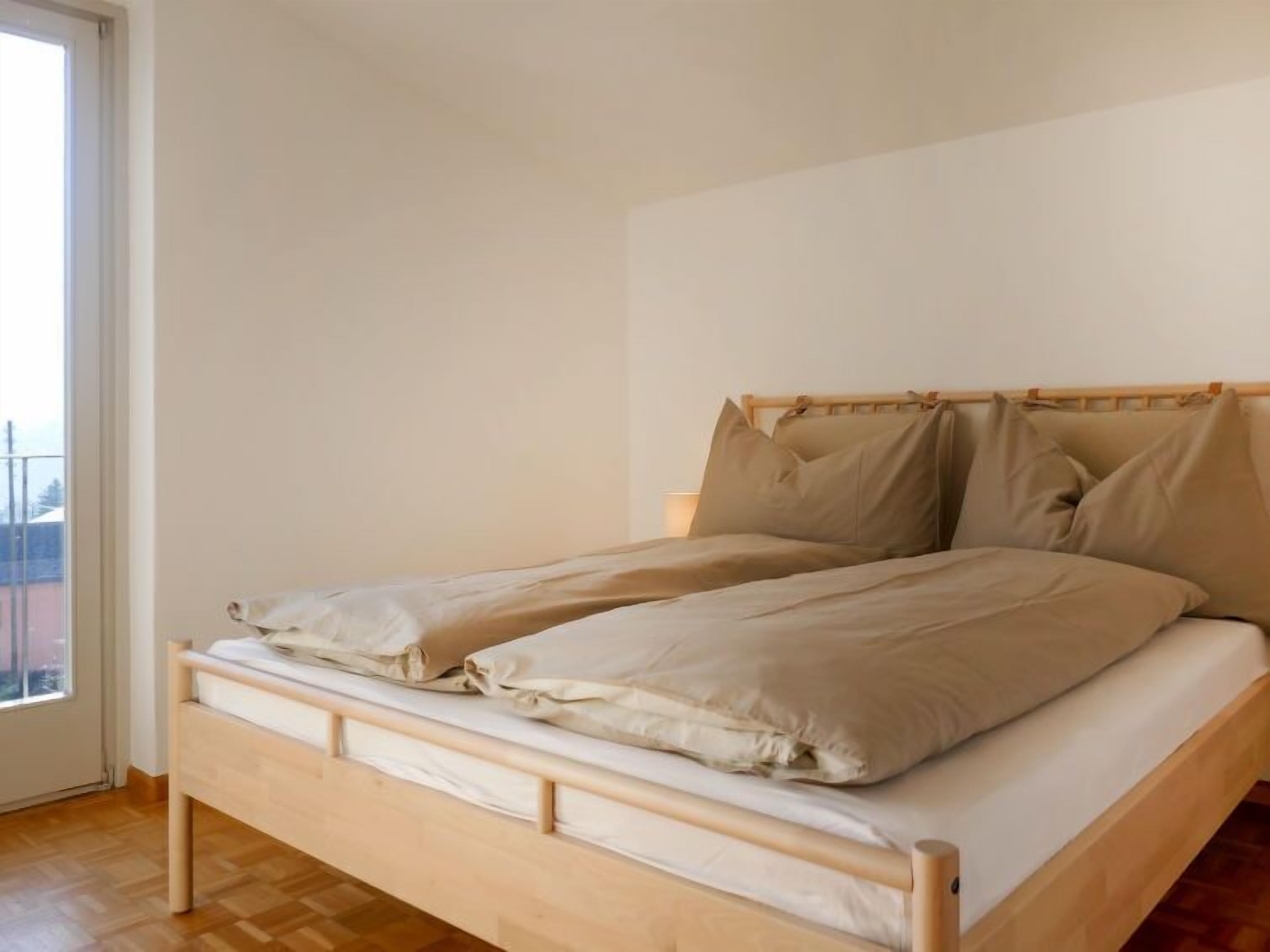 Un lit simple avec un cadre en bois et une literie beige, dans une pièce dotée d’un plancher en bois et d’un mur blanc uni.
