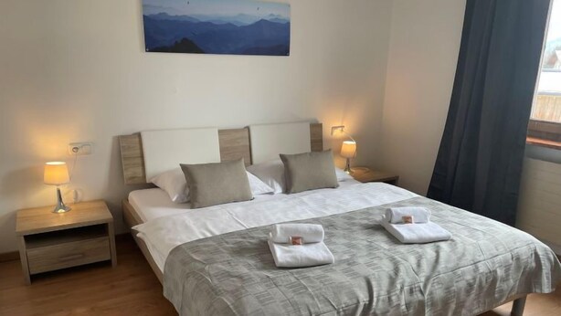 Un letto matrimoniale rifatto con cura, con una coperta grigia, due cuscini e due asciugamani piegati. Sono presenti due comodini con lampade, un quadro appeso al muro e una finestra con tende.