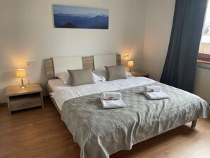 Una cama doble impecablemente vestida con una colcha gris, dos almohadas y dos toallas dobladas sobre la cama. Hay dos mesitas de noche con lámparas, un cuadro en la pared y una ventana con cortinas.