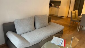 Un soggiorno moderno con un divano grigio, un tavolino da caffè in vetro e una cucina sullo sfondo.