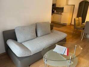 A modern living room with a grey sofa, a glass coffee table, and a kitchen in the background.