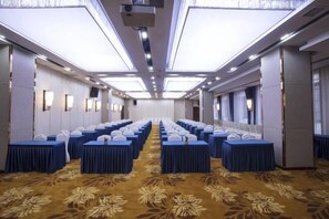 Un salón de conferencias con mesas dispuestas en filas, alfombra estampada y un techo alto con iluminación empotrada.