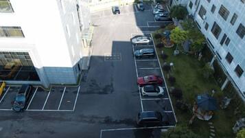 An aerial view of a parking lot with multiple cars, a building with a white facade, and a grassy area with trees and a small structure.