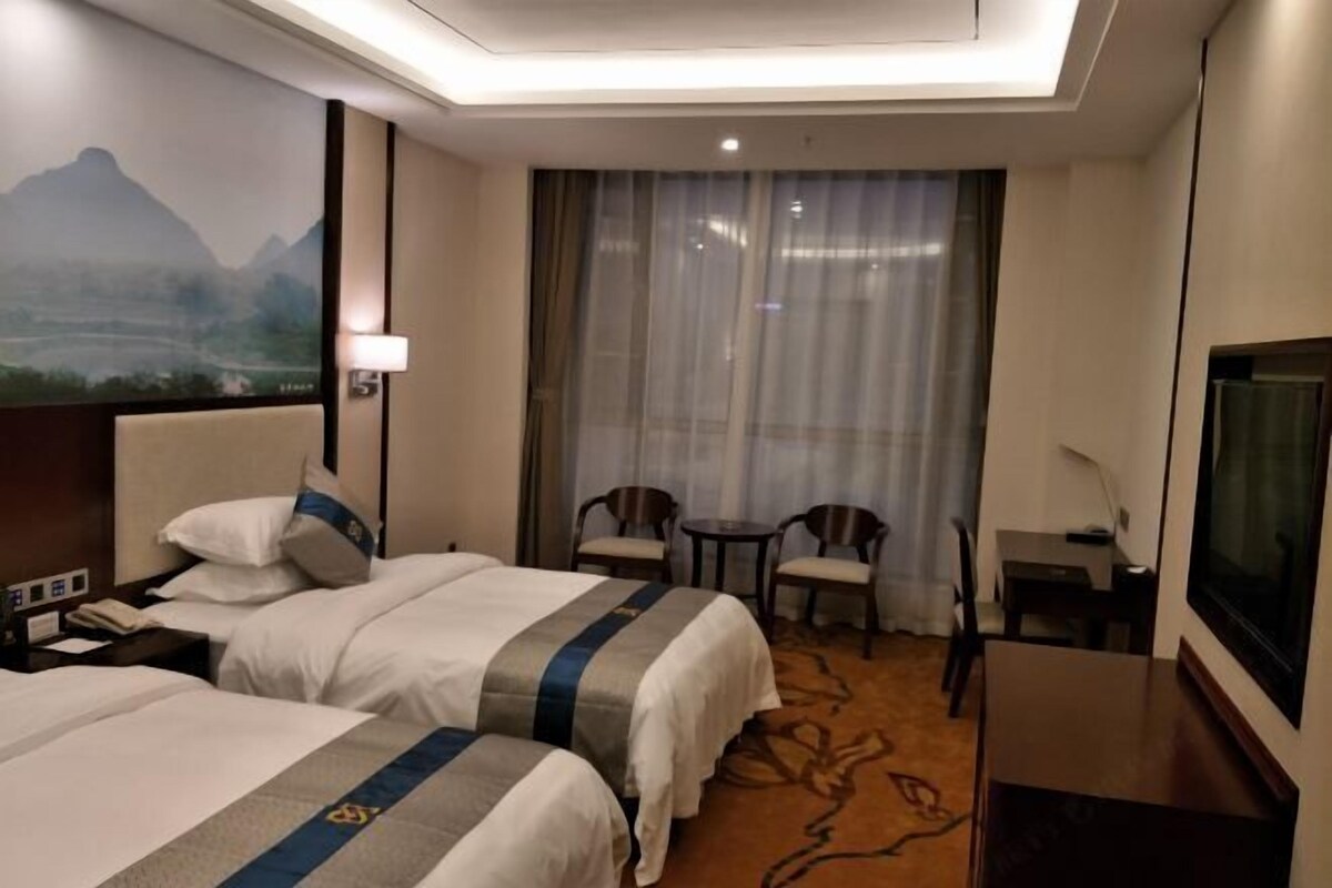 A hotel room with two beds, a desk, a chair, a TV, and a large window with curtains.